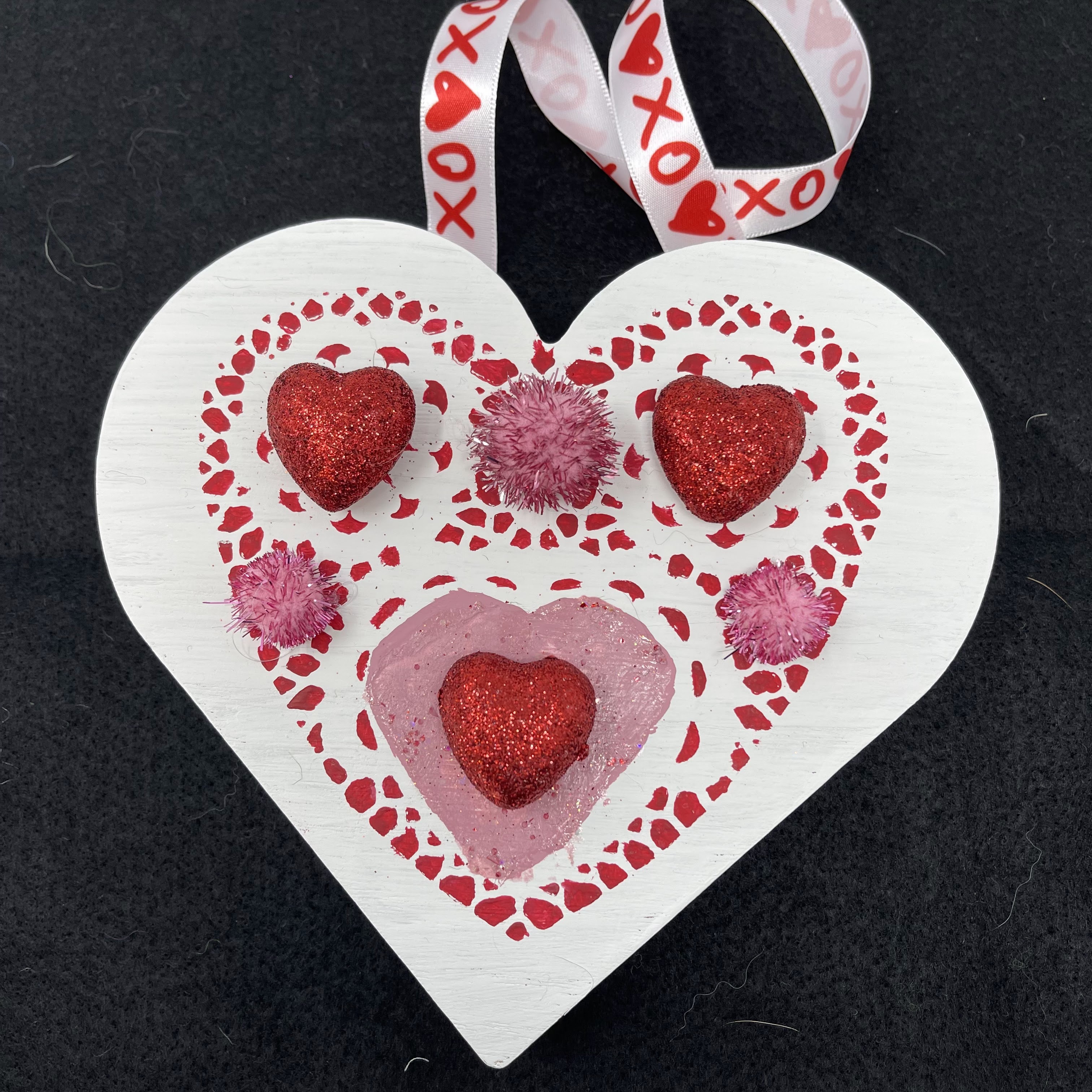 #6 White painted wood heart with red glitter heart and pink pom pom decoration. Hung on white ribbon with red X and O design. 6x7x1"