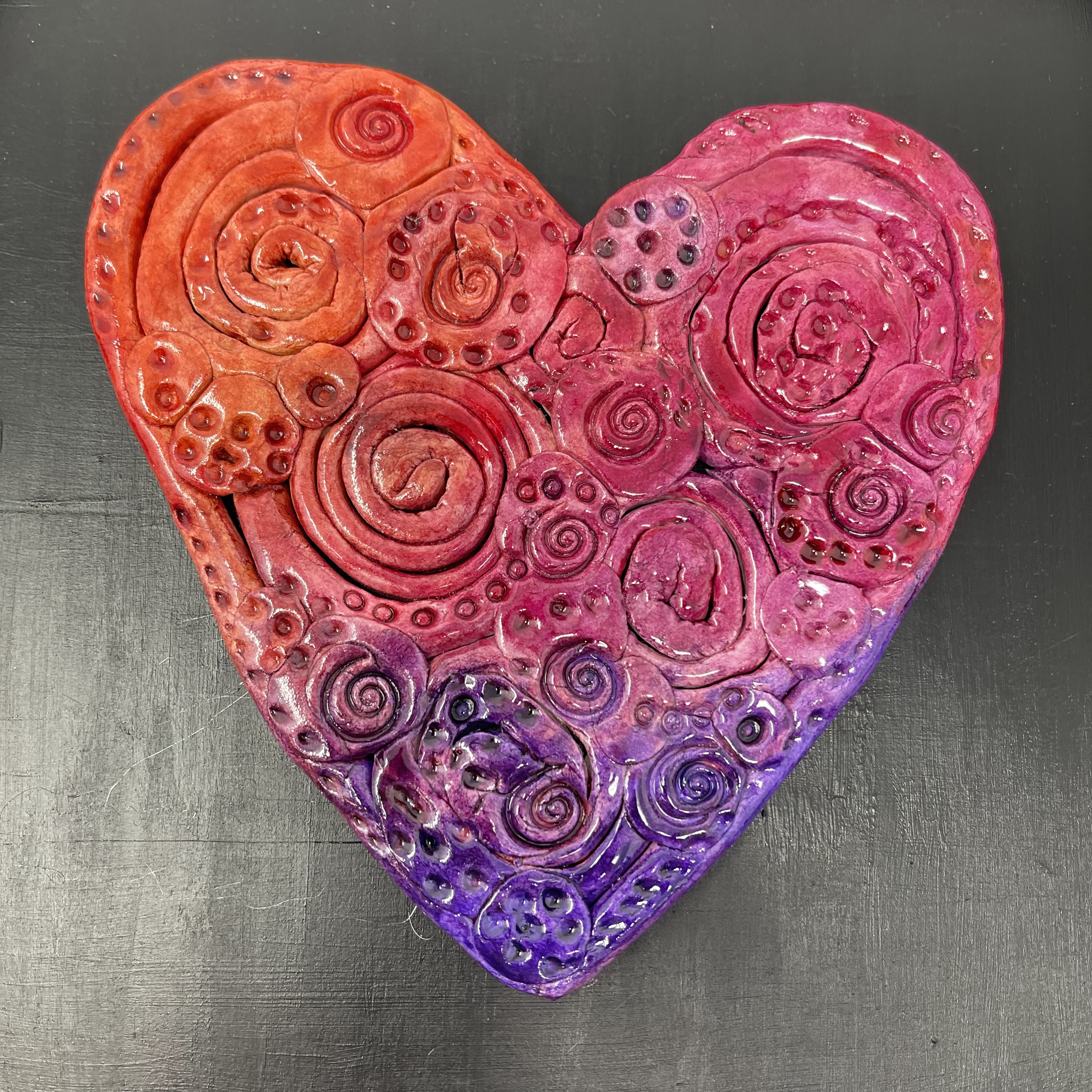 #9 Clay heart decorated with orange, red, and purple spirals. Mounted on black wood frame. 9x9x0.25"